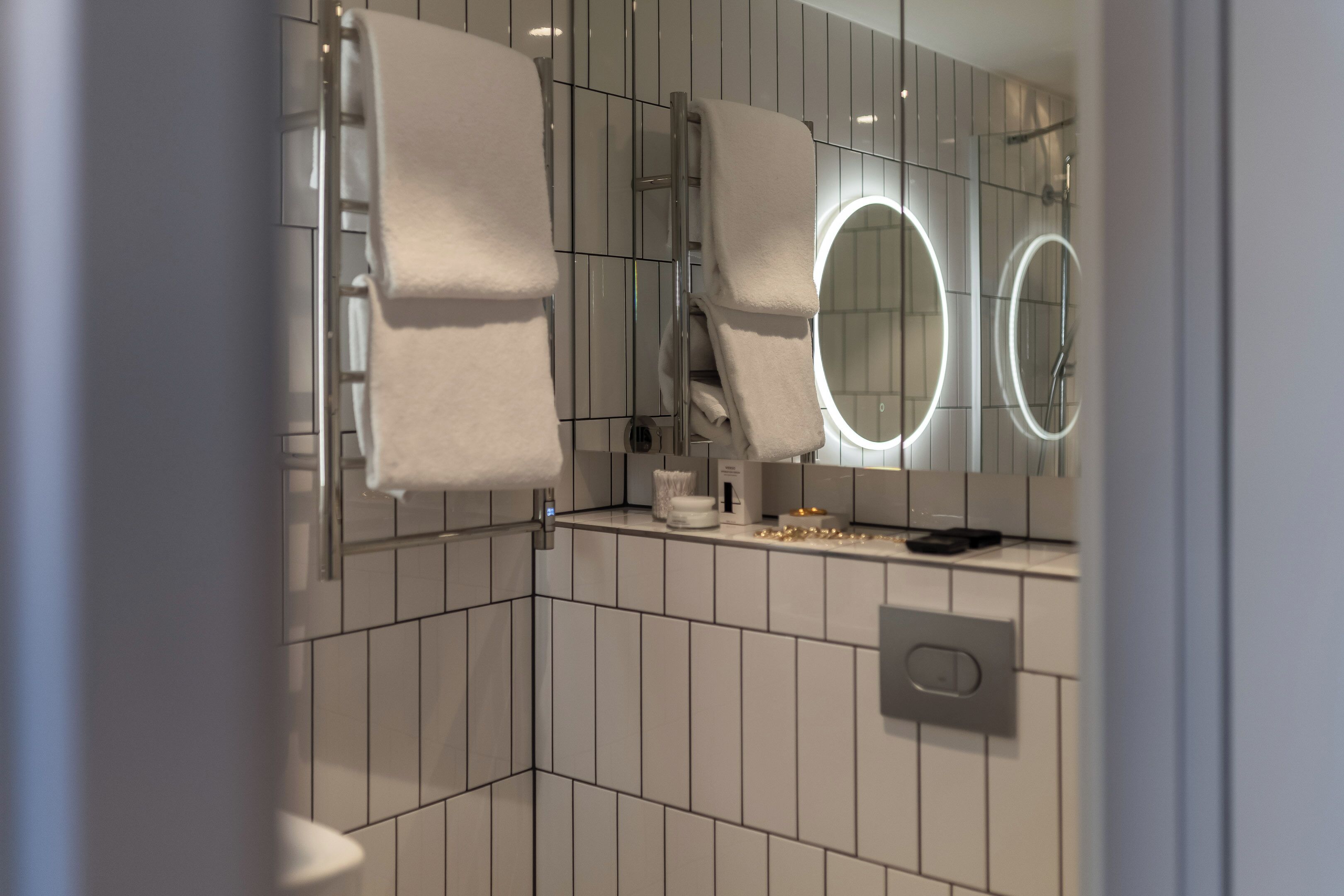 Un baño moderno con azulejos blancos, un perchero, un espejo con luces circulares y una zona de ducha.