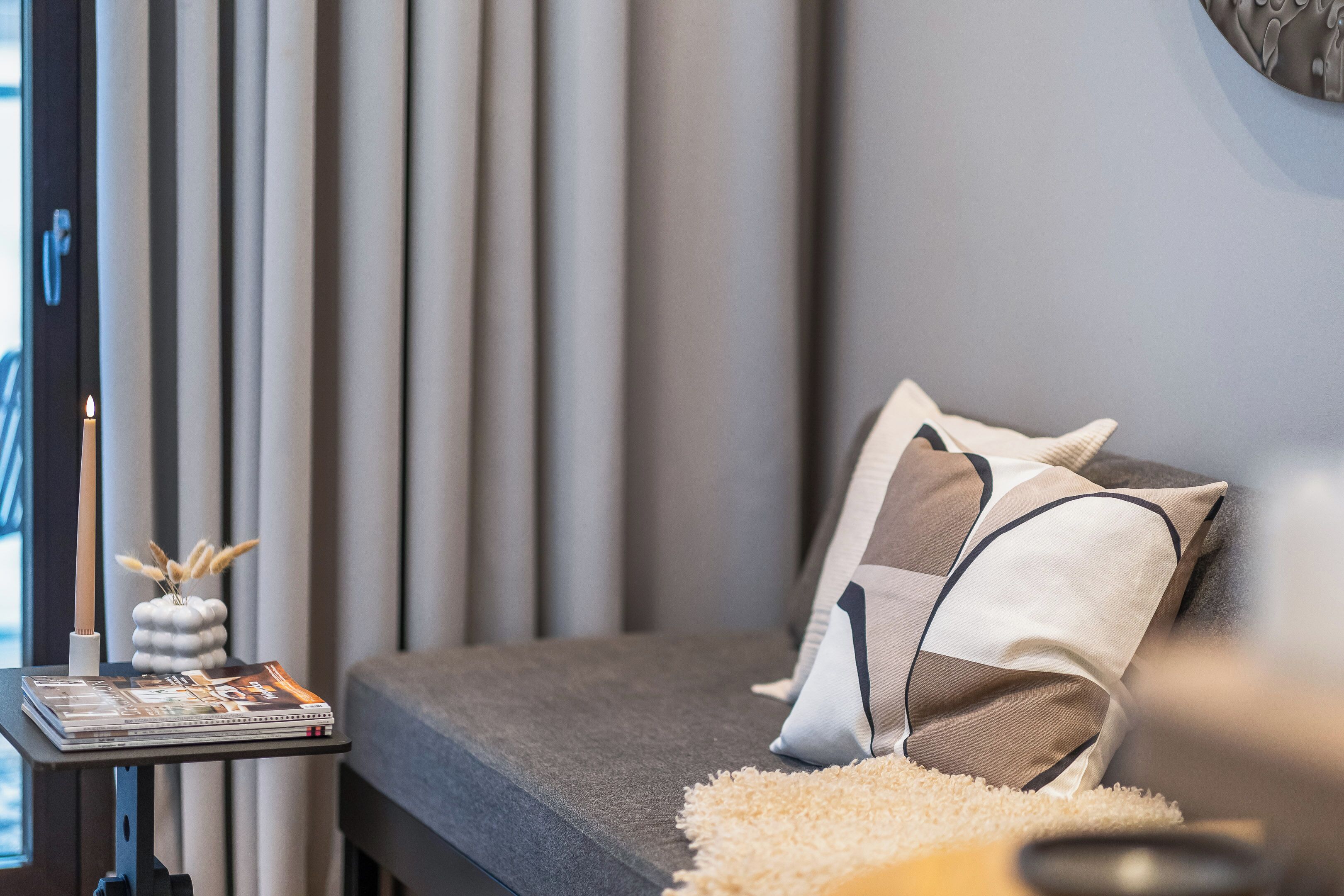 A modern bedroom with a grey sofa, a decorative pillow, a small table with magazines, and a vase with dried plants.