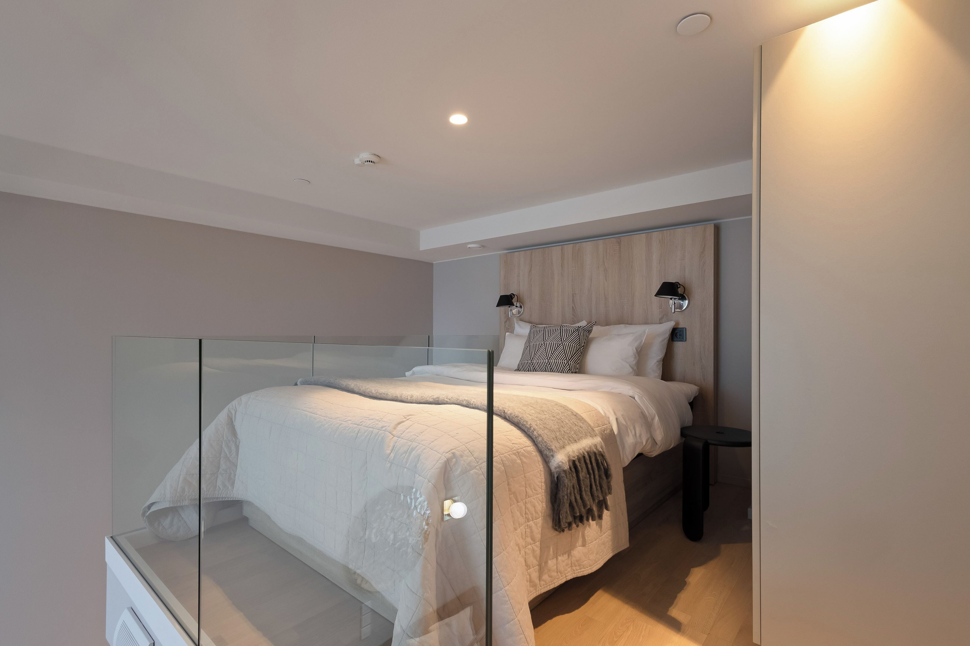 A modern bedroom with a glass partition, a bed with a white duvet, a bedside table, and a nightstand.