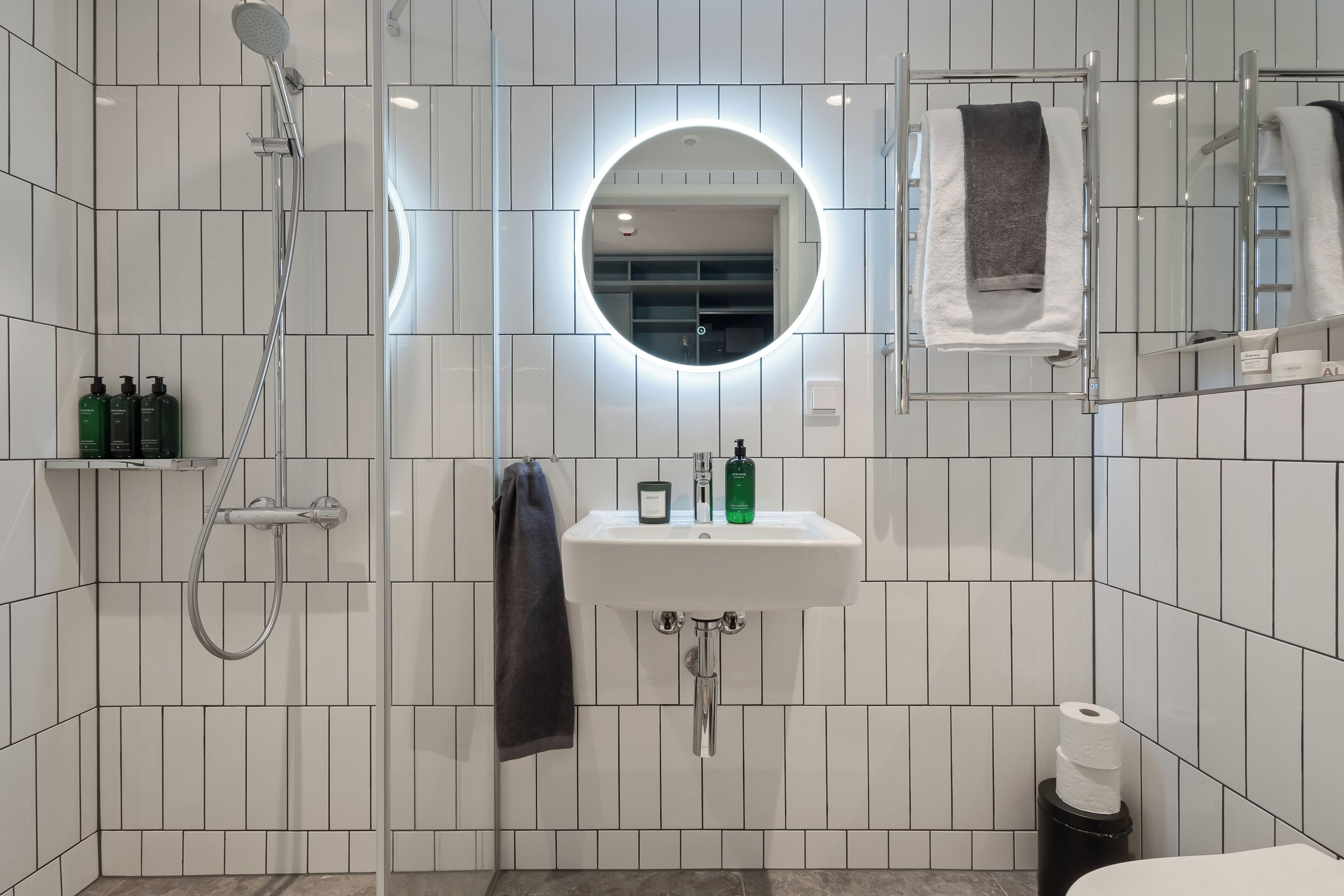 A modern bathroom with a white tiled wall, a round mirror with a light, a white sink, and a shower.