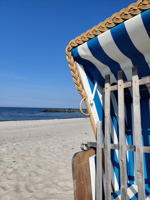 Une chaise de plage recouverte d’une toile à rayures bleues et blanches, avec une bordure tissée, est placée sur une plage de sable. En arrière-plan, on aperçoit un ciel bleu dégagé et une mer calme.