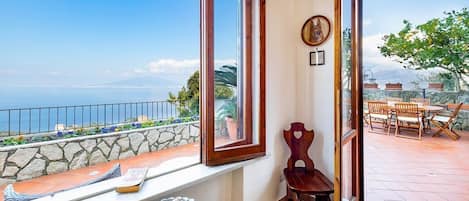 Balcón con vista al mar, pared de piedra, silla de madera y una mesa con una cesta de pan.