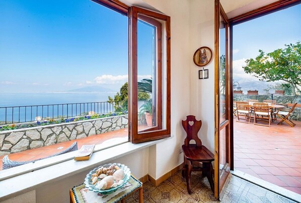 A balcony with a view of the sea, stone wall, wooden chair, and a table with a basket of bread.