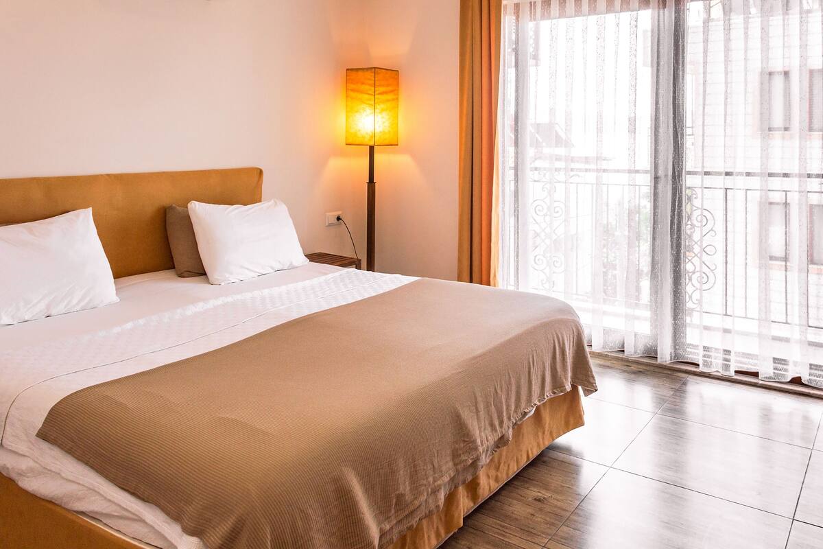 A hotel room with a large bed, a nightstand, a lamp, and a view of the outside through a balcony with curtains.