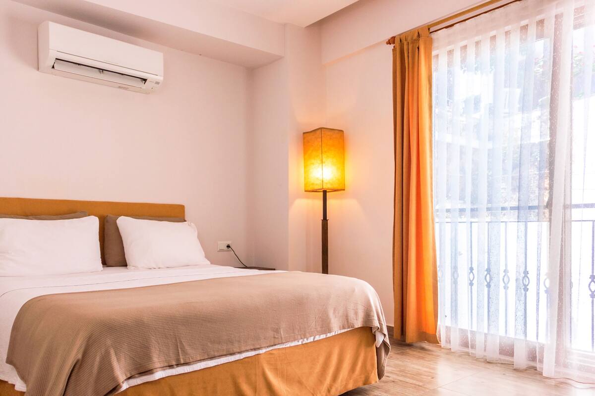 A hotel room with a bed, an air conditioner, a lamp, and a window with curtains.