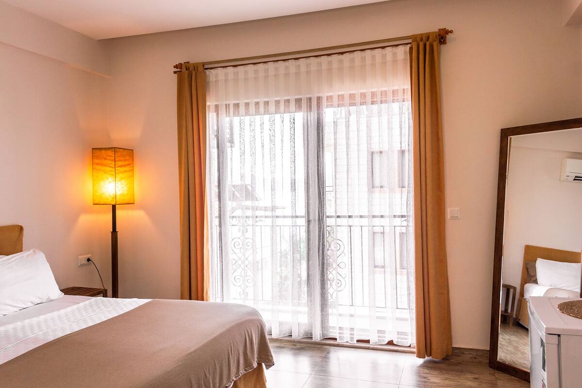 A hotel room with a bed, a nightstand, a lamp, a window with curtains, and a mirror.