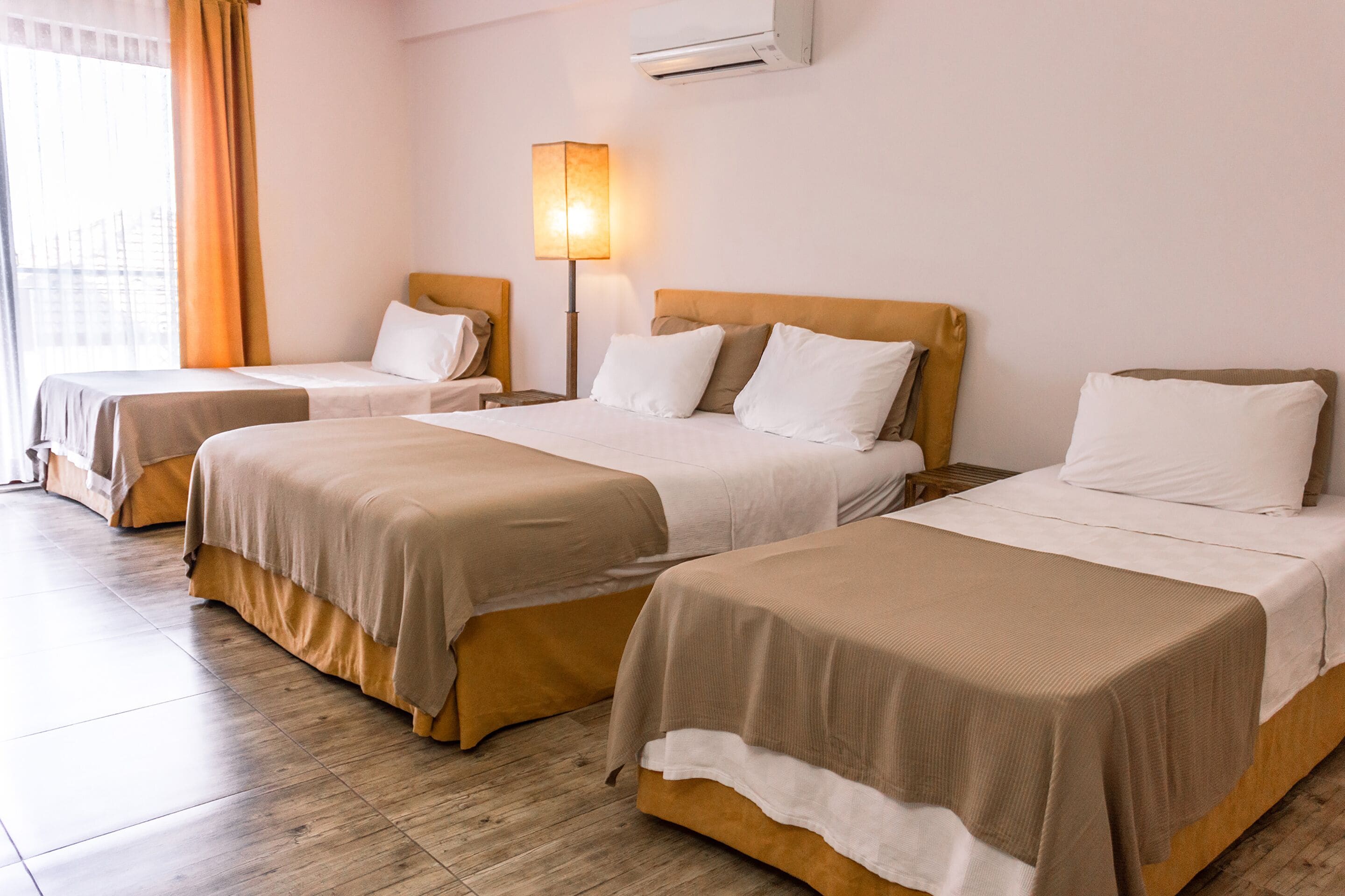 A hotel room with three beds, a nightstand with a lamp, and a window with curtains.