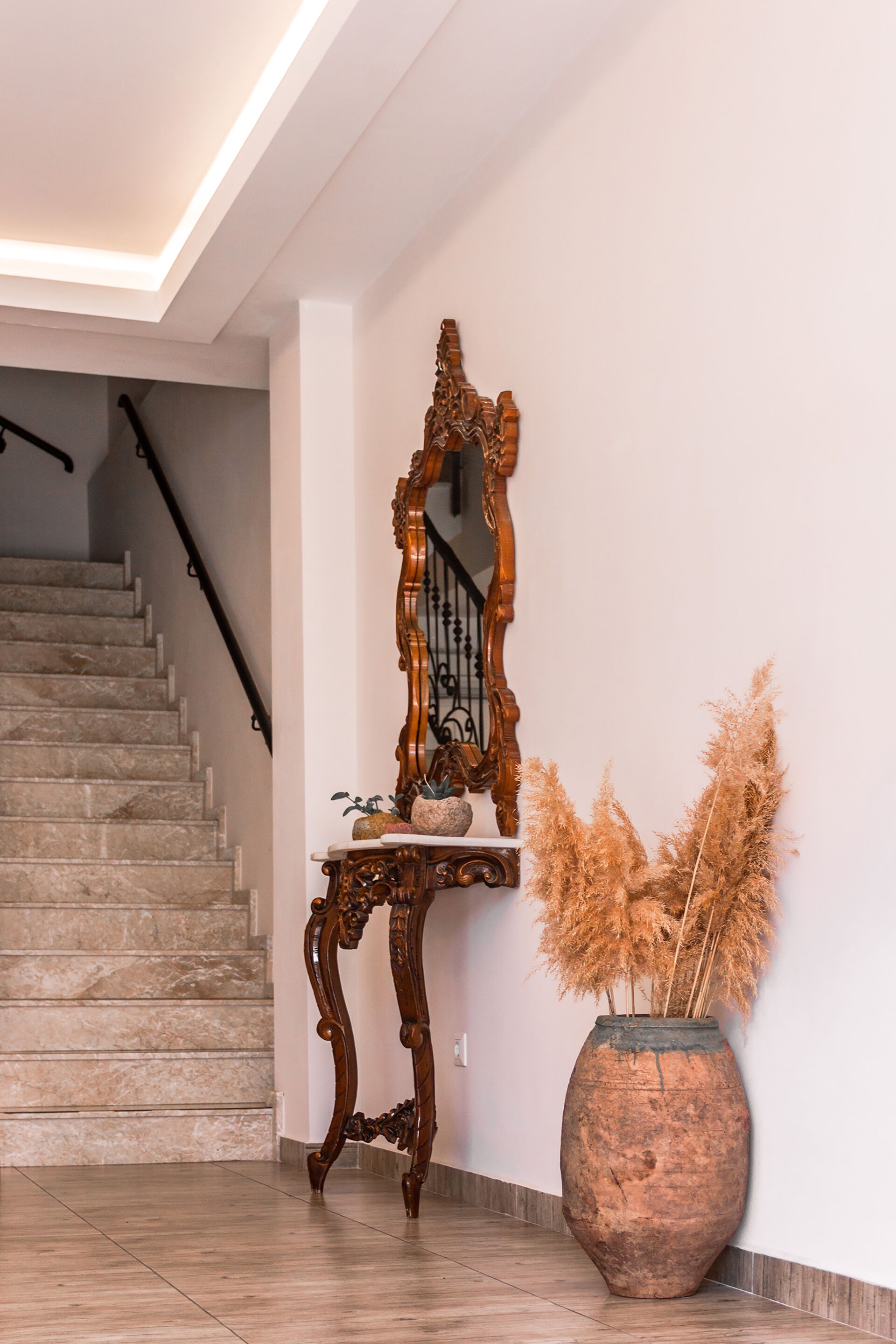 Eine Treppe mit hölzernem Geländer und einer großen Vase mit Pampasgras auf einem Fliesenboden.