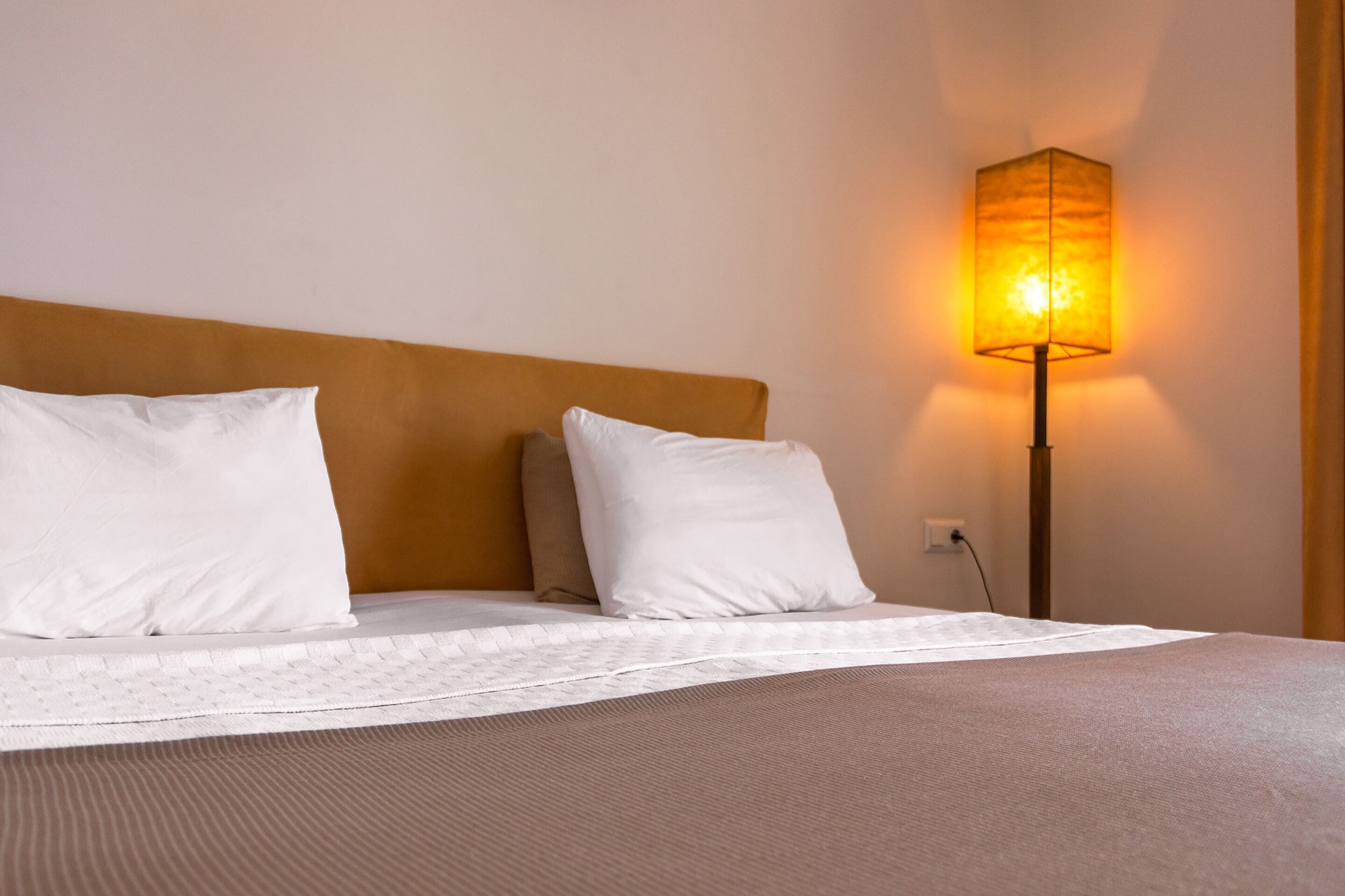 Una cama bien hecha con sábanas blancas y dos almohadas, un cabecero de madera y una lámpara de pie con luz cálida.