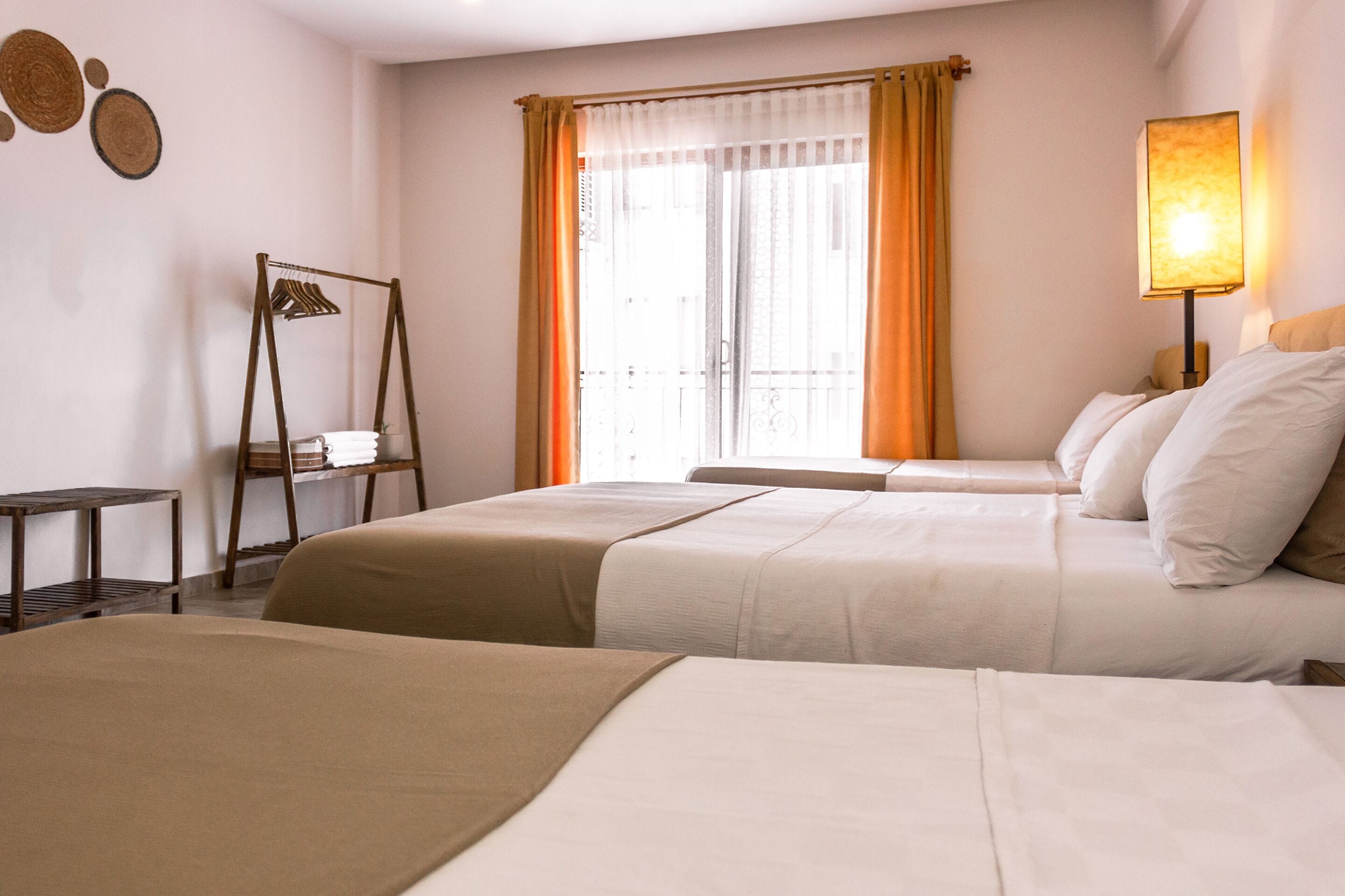 A hotel room with two beds, a small table, a chair, a lamp, and a window with curtains.