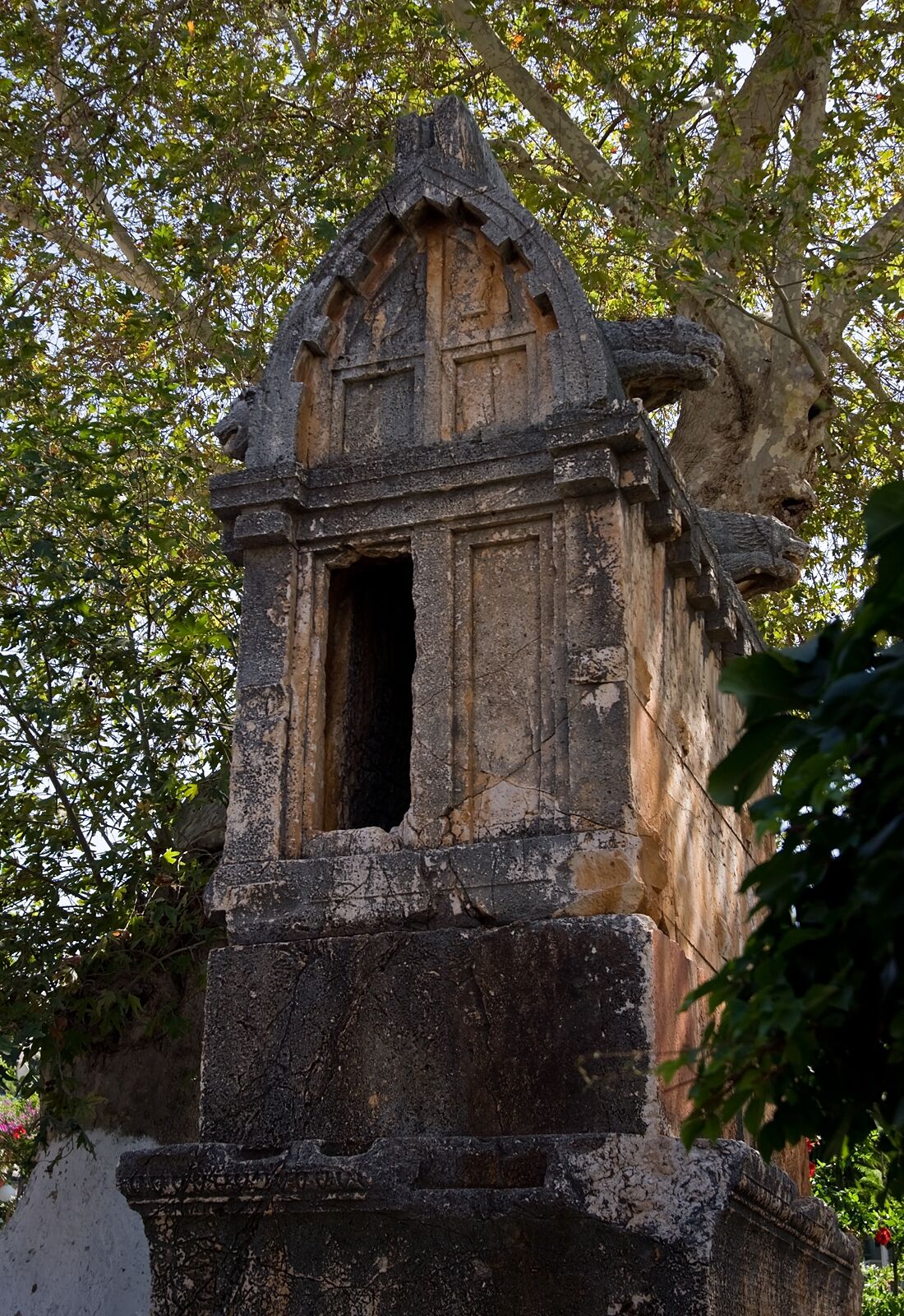A stone structure with a pointed roof and a central opening, surrounded by trees.