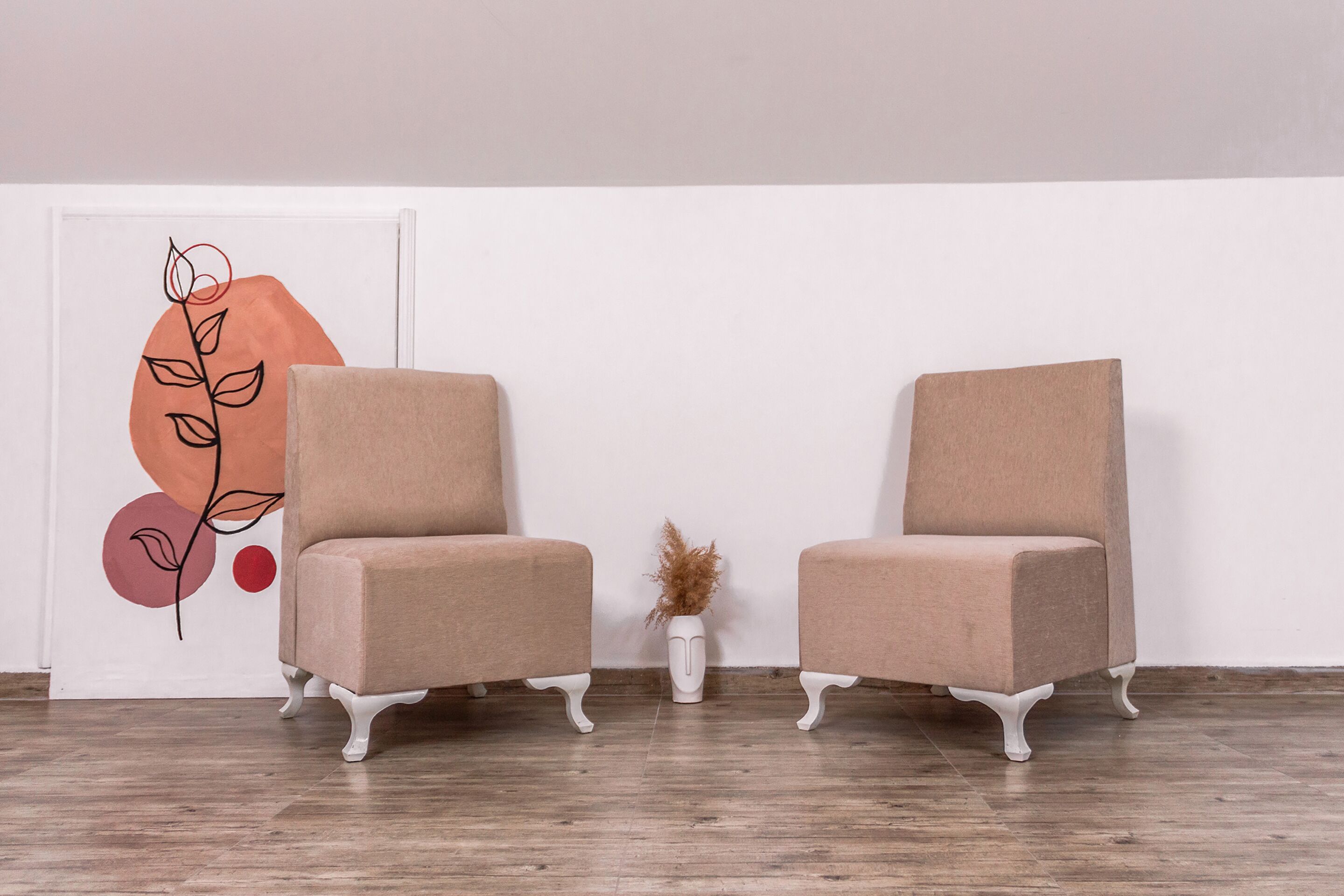 Two beige armchairs with white legs, positioned on a wooden floor against a white wall.