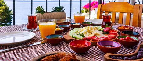 Mesa posta com diversos pratos, incluindo pão, legumes e frutas, com vista para o mar ao fundo.