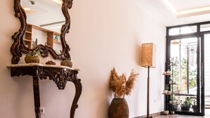 Een hal met een grote, versierde spiegel, een houten console tafel met decoratieve objecten, een hoge vloerlamp en een grote vaas met gedroogde planten.