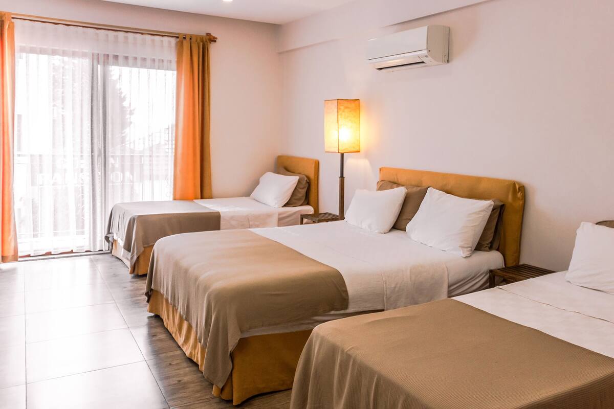 A hotel room with three single beds, a bedside table, a lamp, and an air conditioning unit.