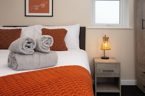 Ein ordentlich bezogenes Bett mit einer weißen und orangefarbenen Decke, zwei zusammengerollten Handtüchern und einer Nachttischlampe auf einem Nachttisch.