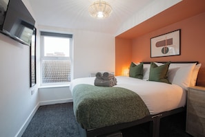 A neatly made bed with a green blanket, a bedside table, a wall-mounted TV, and a window with a view of buildings.