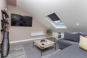 Ein modernes Wohnzimmer mit schrägverdeckter Decke, einem an der Wand montierten Flachbildfernseher, einem grauen Sofa, einem Holztisch und einem Oberlicht.