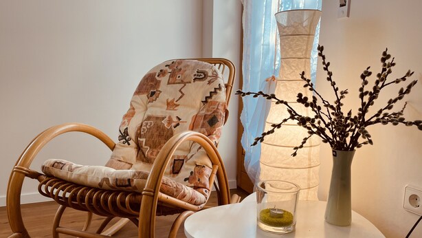 Una poltrona a dondolo in vimini con un cuscino decorato, un tavolino con un vaso di rami e una lampada.