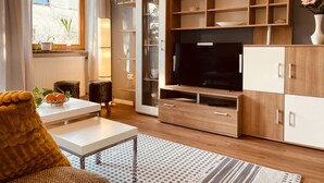 Un soggiorno moderno con televisore a schermo piatto, scaffali in legno, un tavolo bianco e un divano confortevole.