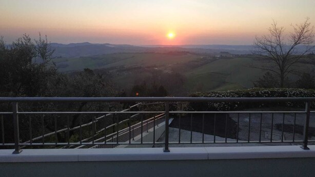 A sunset view from a balcony with a metal railing, overlooking a valley with rolling hills and sparse trees. - Apartment with beautiful view of the hills, between Pesaro and Fano (Mombaroccio)