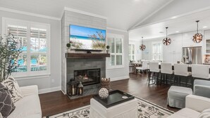 A spacious living room with a fireplace, a large TV, and a dining area with white chairs. - Wood Beach Retreat – Perfect 30A Location offering Easy Beach Access & Private Pool (Santa Rosa Beach)