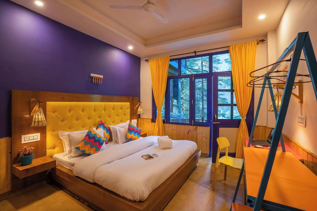 A bedroom with a large bed, a wooden headboard, a bedside table with a lamp, a window with curtains, and a chair.