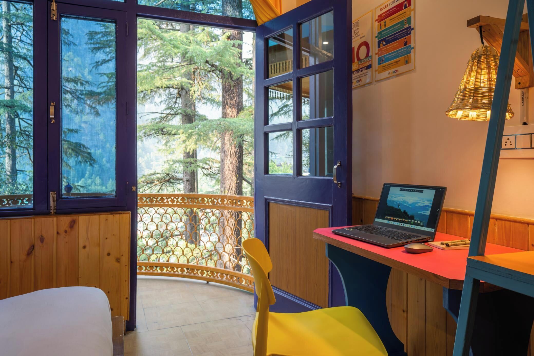 A room with a wooden desk, a laptop, and a chair. There is a large window with a view of trees.