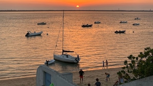 Tramonto su un mare calmo con diverse imbarcazioni ancorate, una spiaggia sabbiosa e alcune persone che passeggiano.