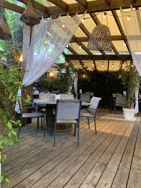 Un espace de restauration en plein air avec des tables et des chaises en bois, des guirlandes lumineuses et une pergola recouverte d’un tissu blanc.
