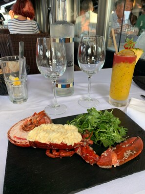 A lobster meal on a black slate board, accompanied by a bottle of water, two wine glasses, and a cocktail with a lemon slice and strawberry garnish.