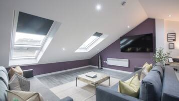 Un soggiorno moderno con soffitto spiovente, lucernari, una televisione a schermo piatto e un divano grigio.
