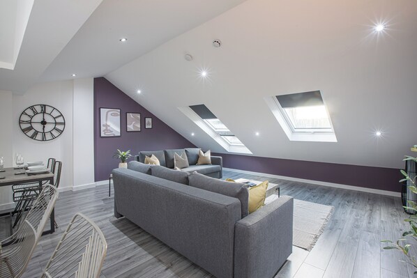 A modern living room with a grey sofa, a dining table with chairs, and a large wall clock.