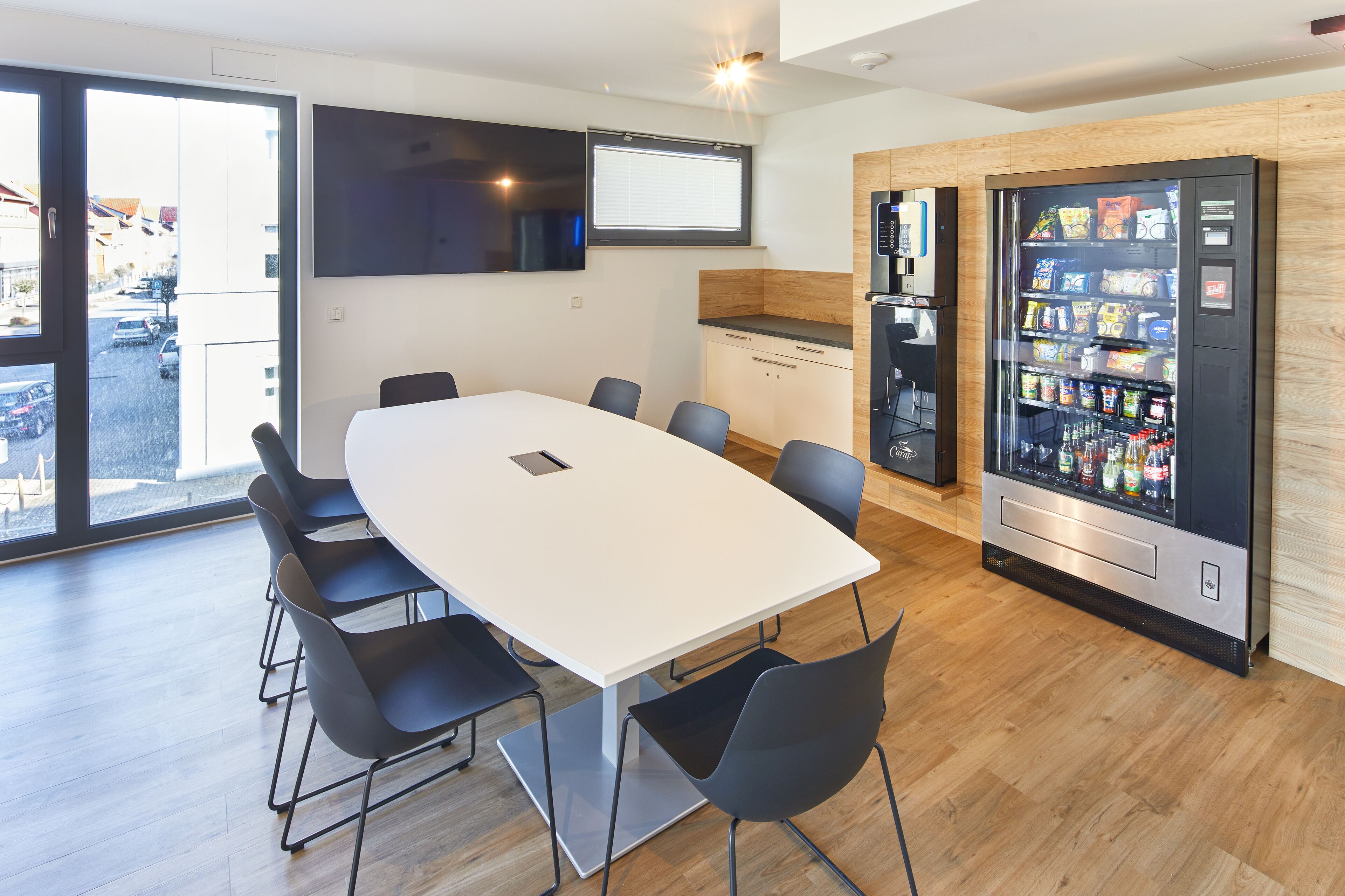 A modern meeting room with a large table, black chairs, a flat-screen TV, a vending machine, and a view of the street through large windows.