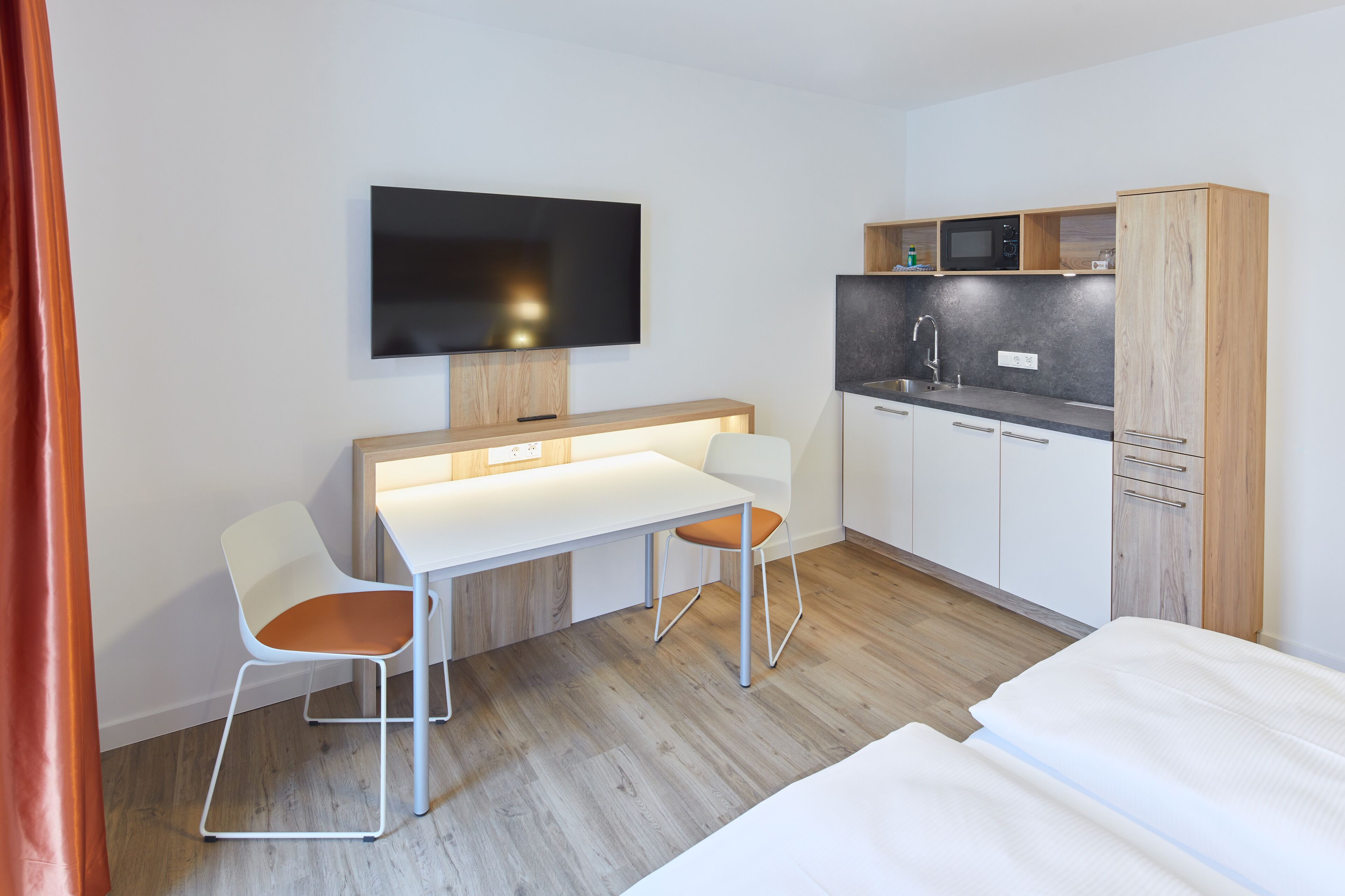 A modern hotel room with a kitchenette, a flat-screen TV, a dining area with two chairs, and a bed.