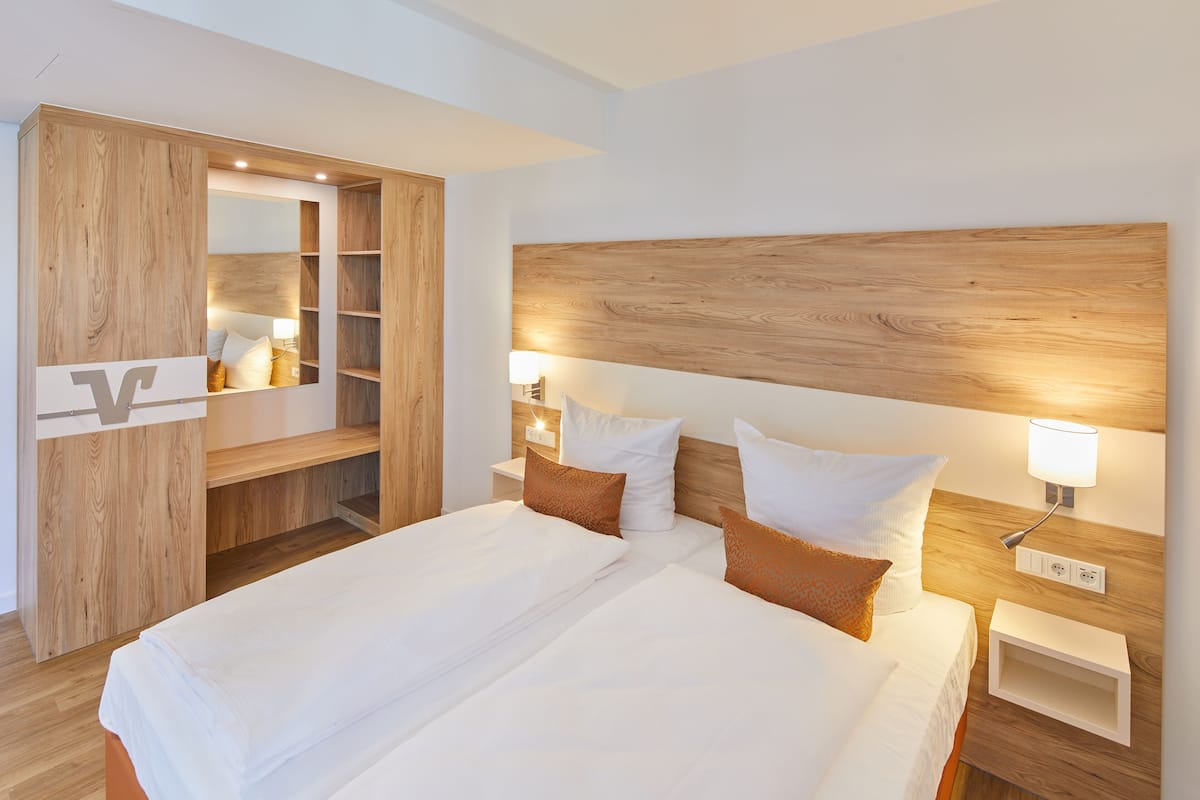 A modern hotel room with a large bed, wooden headboard, bedside lamps, and a built-in wardrobe.