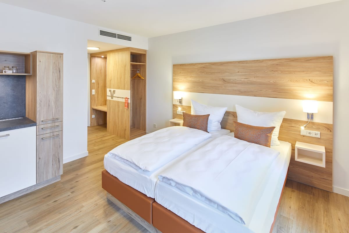 A modern hotel room with a large bed, wooden headboard, bedside tables with lamps, a wardrobe, and a small kitchenette area.