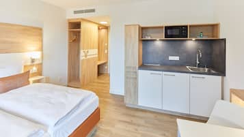 A modern hotel room with a wooden headboard, a bed with white linens, a wooden wardrobe, a kitchenette with a microwave and sink, and a small table.