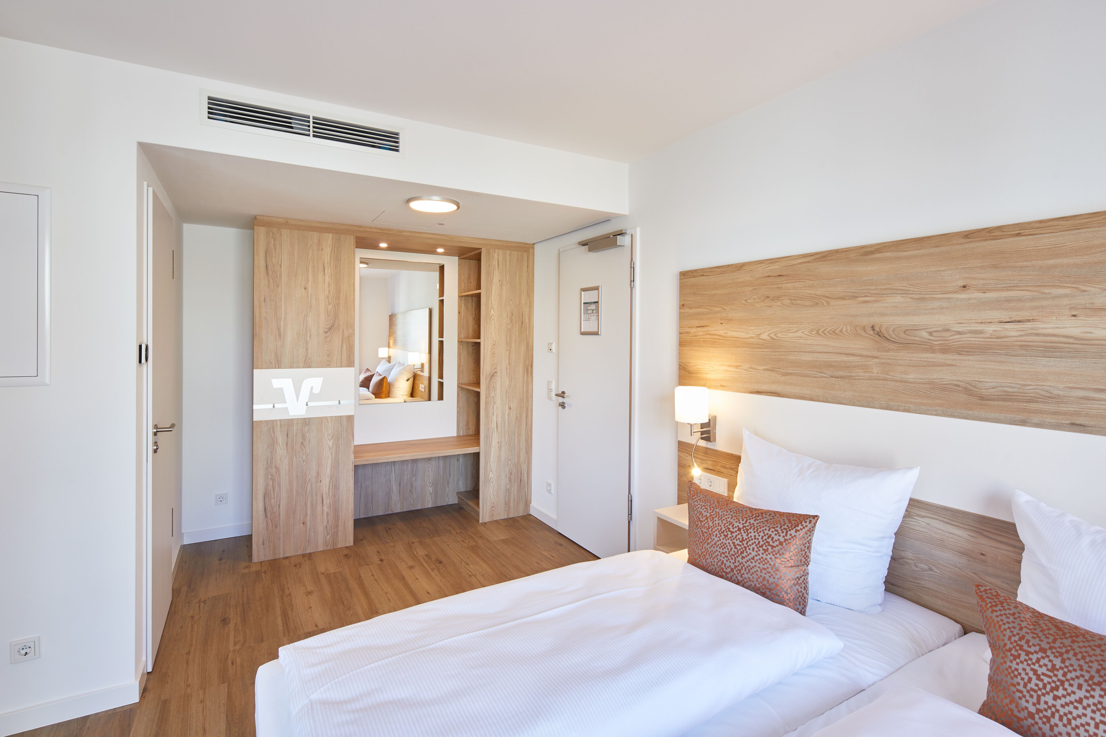 A hotel room with a wooden headboard, a large bed with white bedding, a bedside table with a lamp, and a wooden wardrobe.