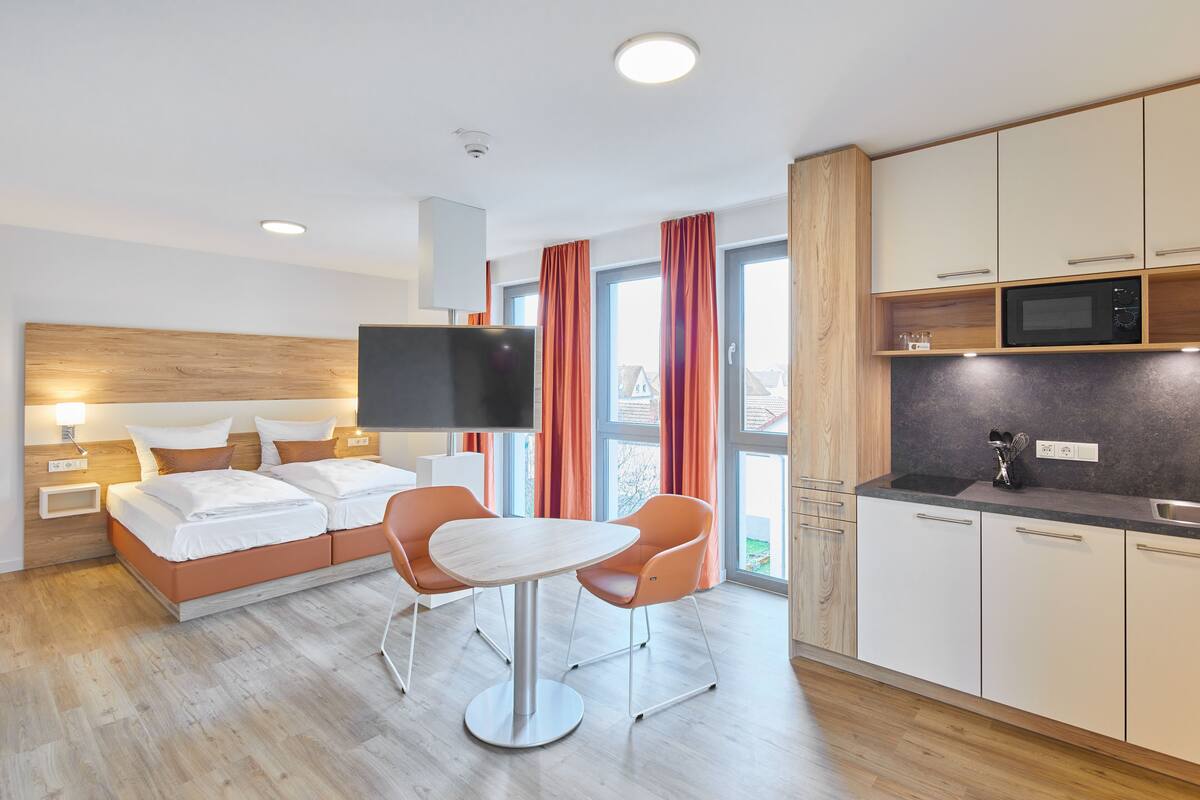 A modern hotel room with a bed, a TV, a dining table, and a kitchen area.