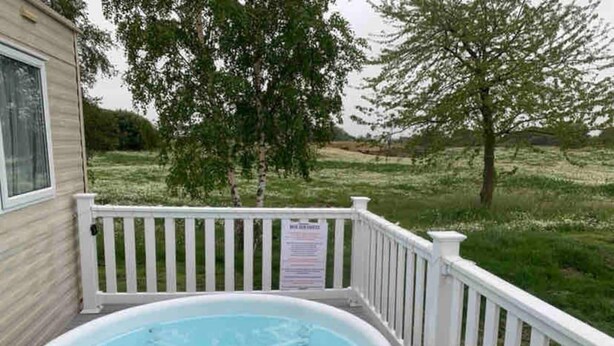 A hot tub on a wooden deck with a white railing, surrounded by a grassy yard and trees.