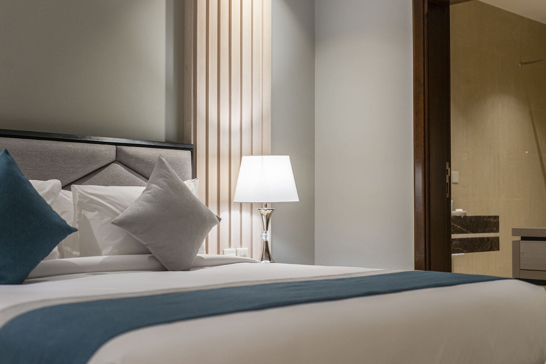A neatly made bed with a blue throw blanket, a white pillow, and a blue pillow. There is a bedside lamp with a white shade.