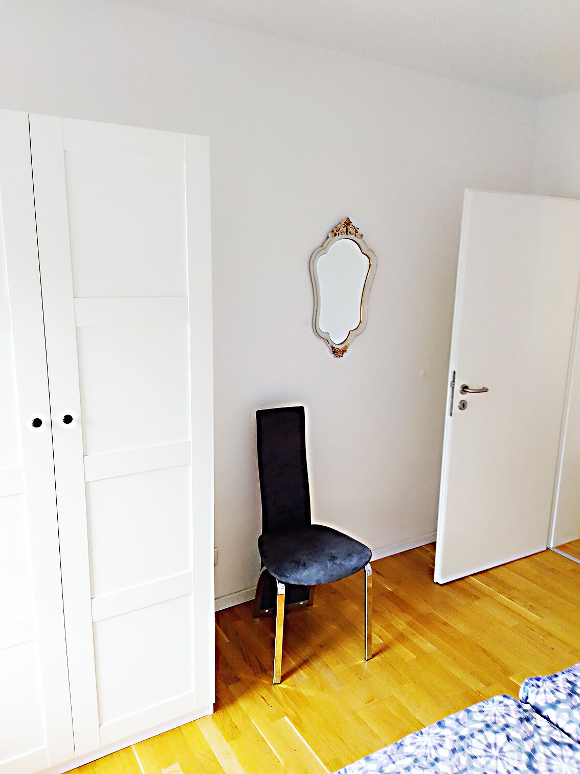 A room with a wooden floor, a black chair, a white door, and a mirror on the wall.