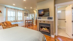A bedroom with a fireplace, a television mounted above it, a bed, a sofa, and a small dining area with a table and chairs.