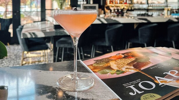 A bar area with a cocktail, a magazine, and a TV screen in the background.