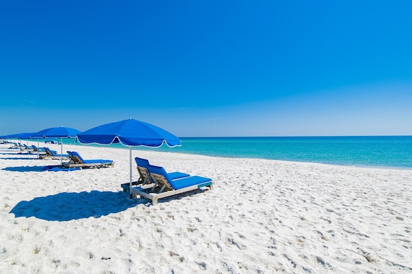 A beach with white sand and clear blue water. There are several blue umbrellas and lounge chairs.