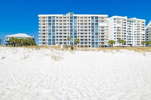 A beachfront hotel with multiple floors, a sandy beach in the foreground, and palm trees.