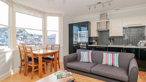 A modern living room with a grey sofa, wooden dining table, and chairs. There is a kitchen area with black cabinets and a window overlooking a cityscape. - River View (Dartmouth)