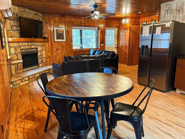 Un intérieur en bois avec une cheminée en pierre, une table à manger ronde entourée de chaises, un canapé en cuir noir et un réfrigérateur.