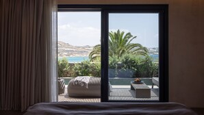 A bedroom with a large sliding glass door leading to a balcony with a view of the sea and greenery. - Noima Boutique Hotel Mykonos (Mykonos)